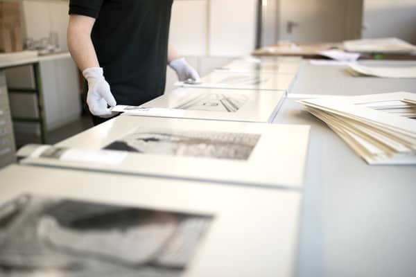 Rahmung diverser Ausstellungsexponate durch Mitarbeiter mit behandschuhten Händen. Die Exponate liegen nebeneinander auf einer grauen Arbeitsfläche