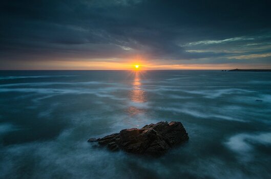 Langzeitbelichtung am Meer mit Felsen im Vordergrund und Sonnenuntergang über dem Ozean