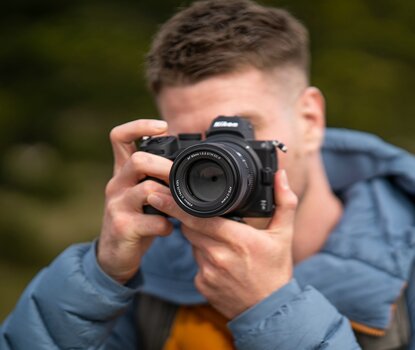 Fotograf blickt durch den Sucher einer spiegellosen Systemkamera mit montiertem Viltrox AF 90mm F2.2 EVO Teleobjektiv. Fotograf blickt durch den Sucher einer spiegellosen Systemkamera mit montiertem Viltrox AF 90mm F2.2 EVO Teleobjektiv.