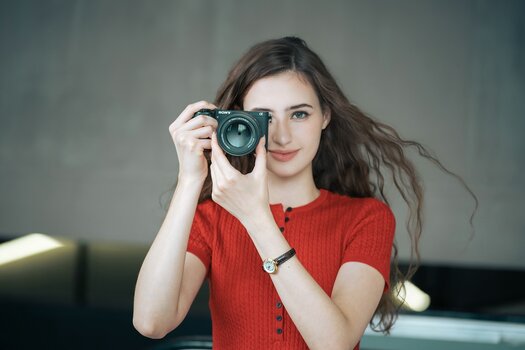 Frontalansicht einer lächelnden Frau mit braunen, welligen Haaren im roten Oberteil, die eine Sony Alpha Kamera mit dem Viltrox AF 75mm F1.8 EVO vor ihr rechtes Auge hält. Frontalansicht einer lächelnden Frau mit braunen, welligen Haaren im roten Oberteil, die eine Sony Alpha Kamera mit dem Viltrox AF 75mm F1.8 EVO vor ihr rechtes Auge hält.