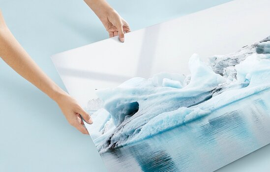Nahaufnahme von zwei Händen, die ein glänzendes, großformatiges Acrylglas-Wandbild mit einer Eisberglandschaft anheben. Nahaufnahme von zwei Händen, die ein glänzendes, großformatiges Acrylglas-Wandbild mit einer Eisberglandschaft anheben.