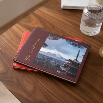 Zwei geschlossene Fotobücher liegen übereinander auf einem Holztisch, daneben steht ein Wasserglas.