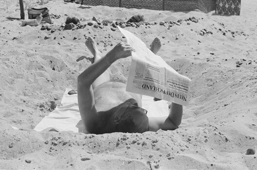 Schwarz-Weiß-Szene aus dem Dokumentarfilm „Hiddensee“: Ein Mann liegt am Strand im Sand und liest eine Zeitung.