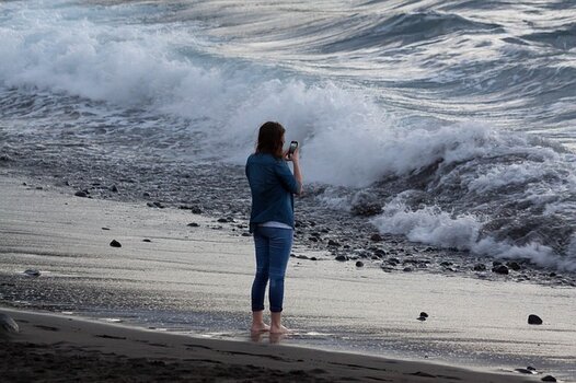 Frau am Strand fotografiert Gischt mit dem Smartphone