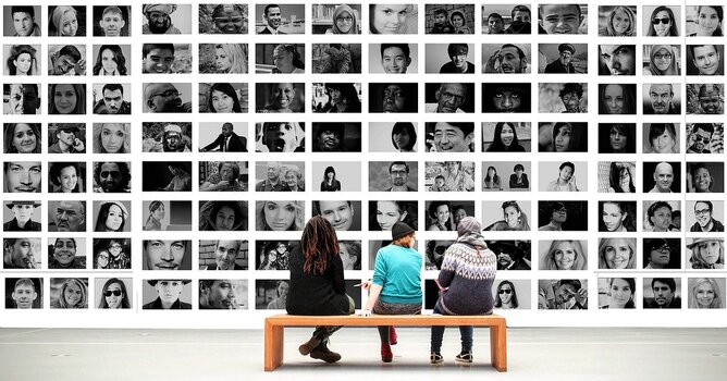 Drei Menschen, auf Holzbank sitzend,  betrachten Wand mit Fotocollage von Portraits 