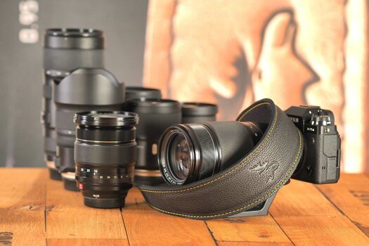 Fujifilm XT4 mit Objektiv und EDDYCAM Kameragurt schwarz mit gelber Kontrastnaht auf Holztisch, dahinter stehen mehrere Objektive