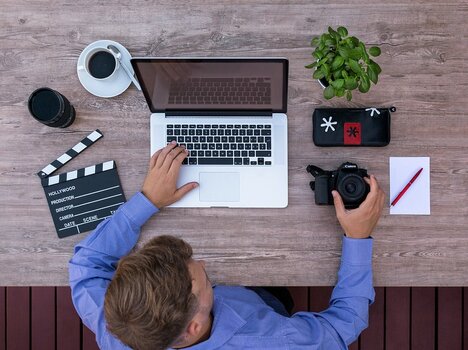 Blick von oben: Mann am Tisch vor Laptop, Kamera und Büroutensilien