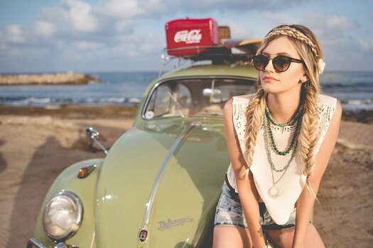 Hippie-Model lehnt an altem VW Käfer mit Coca-Cola-Kühlbox auf dem Dach. Szene am Strand.