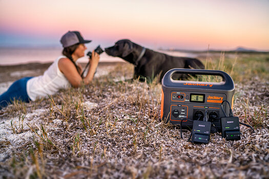 Frau fotografiert liegenden Hund auf Augenhöhe. Im Vordergrund Jackery Explorer 1000 mit zwei Ladegeräten für Sony-Akkus