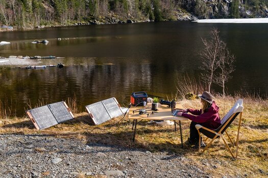 Fotografin an einem See arbeitet an ihrem Laptop auf einem Campingtisch umgeben von Kamera und Drohne. Alle Geräte werden mit dem Jackery Solargenerator und 2 Solarpanels aufgeladen.