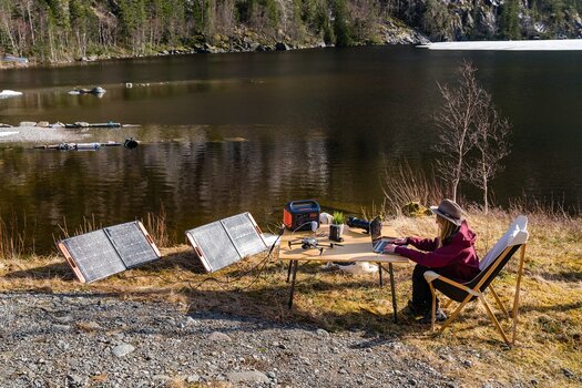 Fotografin an einem See arbeitet an ihrem Laptop auf einem Campingtisch umgeben von Kamera und Drohne. Alle Geräte werden mit dem Jackery Solargenerator und 2 Solarpanels aufgeladen. Fotografin an einem See arbeitet an ihrem Laptop auf einem Campingtisch umgeben von Kamera und Drohne. Alle Geräte werden mit dem Jackery Solargenerator und 2 Solarpanels aufgeladen.