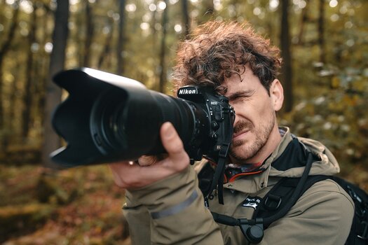 Mann fotografiert mit Nikon D810, Kamera ist mit dem SNAPSNAP camera strap von FIDLOCK am Rucksack befestigt Mann fotografiert mit Nikon D810, Kamera ist mit dem SNAPSNAP camera strap von FIDLOCK am Rucksack befestigt