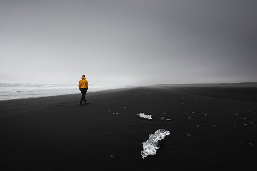 Person mit orangefarbener Jacke geht am dunklen Meeresstrand entlang. Auf dem Sand liegen bizarre Eisformationen.