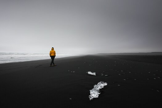 Person mit orangefarbener Jacke geht am dunklen Meeresstrand entlang. Auf dem Sand liegen bizarre Eisformationen. Person mit orangefarbener Jacke geht am dunklen Meeresstrand entlang. Auf dem Sand liegen bizarre Eisformationen.