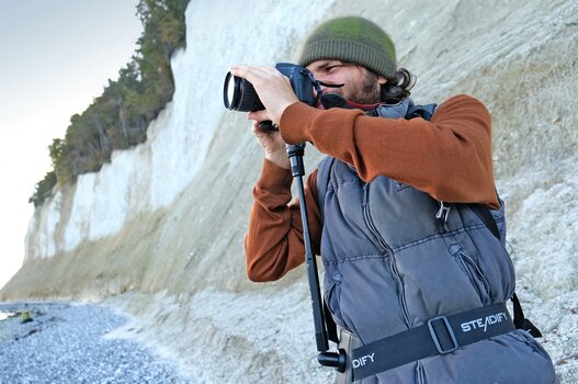 Mann mit Weste und Mütze fotografiert am Fuß einer Steilküste, die Kamera ist auf dem Hüftstativ Steadify montiert. 