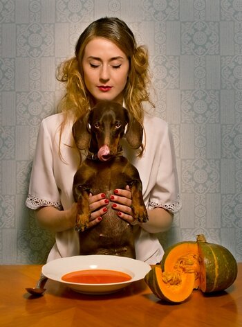 Frau mit geschlossenen Augen sitzt an Tisch mit gefülltem Suppenteller, daneben ein angeschnittener Kürbis. Auf dem Schoß hält sie aufrecht sitzend einen Hund, der sich mit der Zunge über die Nase leckt.