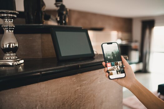 im Wohnzimmer auf einem Kaminsockel stehender schwarzer digitaler Bilderrahmen, davor hält eine Frau ein Smarthphone, um den Bilderrahmen via App Informationen zukommen zu lassen.
