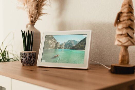 auf einer hellbraunen Kommode steht ein Blumentopf mit Pflanze, daneben ein weißer digitaler Bilderrahmen mit einem Bild, das ein kleines Ruderbot mit einer Person auf einem smaragdgrünen See inmitten von Felswänden abbildet. auf einer hellbraunen Kommode steht ein Blumentopf mit Pflanze, daneben ein weißer digitaler Bilderrahmen mit einem Bild, das ein kleines Ruderbot mit einer Person auf einem smaragdgrünen See inmitten von Felswänden abbildet.