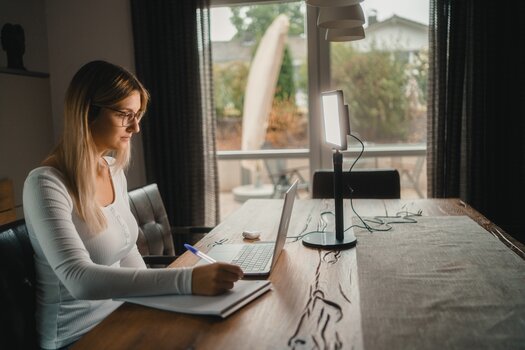 Frau sitzt in Wohnumgebung an einem Tisch vor einem Laptop und macht sich Notizen. Links neben dem Laptop das Lumis Key Pro Light