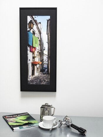 Bunte Straßenszene im Hochformat mit schwarzem Passepartout und Rahmen hängt an einer hellen Wand.