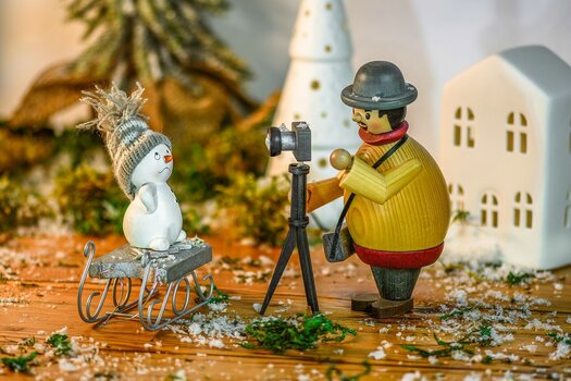 Aus Holzfiguten gebaute Szene: Mann mit Kamera auf Stativ fotografiert Schneemann, der auf einem Schlitten steht.