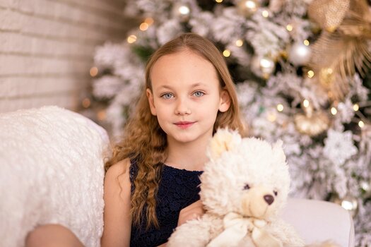 Langhaariges Mädchen mit Stofftier im Arm sitzt auf einem weißen Sofa, hinter ihr ein weißgeschmückter, beleuchteter Weihnachtsbaum.