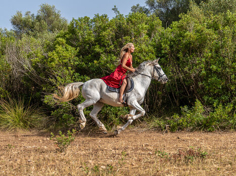 Frau in rotem Sommerkleid reitet auf weißem Pferd von links nach rechts durch den Bildausschnitt.