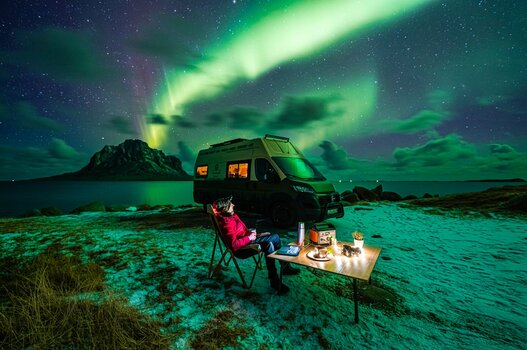 Camper an gedecktem Tisch vor Wohnmobil in nordischer Winterlandschaft mit Polarlicht.