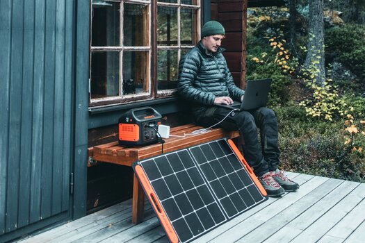 Mann in Winterkleidung sitzt auf der Bank vor einem Holzhaus und bedient seinen Laptop. Neben ihm ein Solargenerator und ein Solarpanel von Jackery.