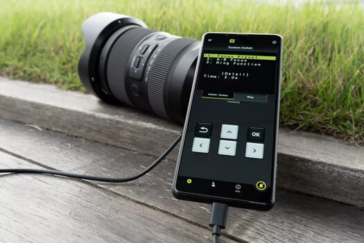 Tamron Objektiv wird mit der Lens Utility App und Smartphone eingestellt. Objektiv und Smartphone liegen auf einem Holzbohlenweg am Rande einer Graswiese.