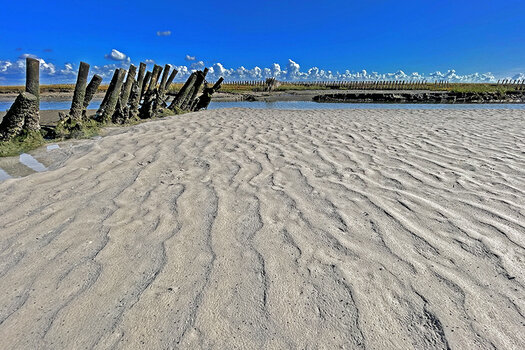 Horizont weit oben im Bild: im Vordergrund fester Sand, links Holzpfähle, im Hintergrund Wolken entlang der Horizontlinie.