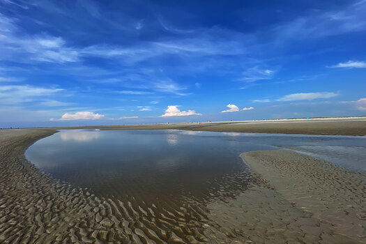 blauer Himmel mit wenigen Wolken, im Vordergrund Wasser und Sandboden 