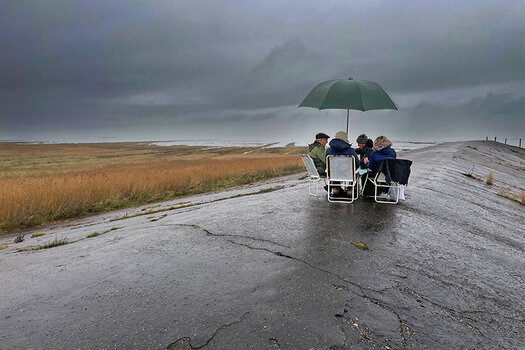 Vier Menschen sitzen bei Regenwetter auf Klappstühlen unter einem großen Schirm