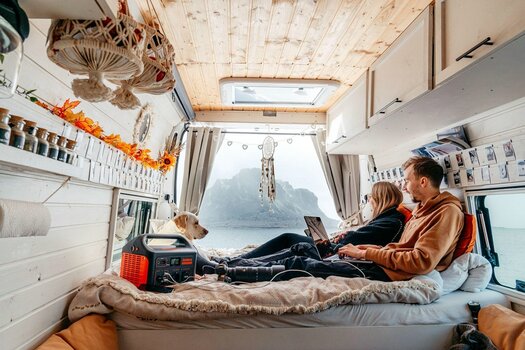 Paar mit Hund sitzt im Wohnwagen oder Wohnmobil auf dem Bett und bedient Laptop und Handy. Die Geräte werden durch einen Jackery Explorer geladen. Paar mit Hund sitzt im Wohnwagen oder Wohnmobil auf dem Bett und bedient Laptop und Handy. Die Geräte werden durch einen Jackery Explorer geladen.