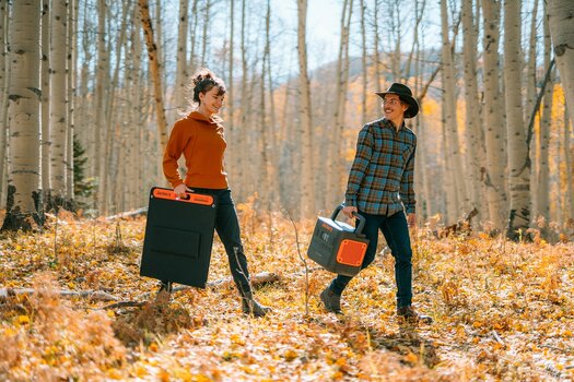 Ein Pärchen geht durch einen sonnigen Herbstwald. Sie trägt ein zusammengeklapptes Solarpanel, er den neuen Jackery 1500 Pro.