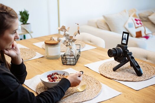 Frau sitzt an Tisch und isst Müsli. Vor ihr die Canon EOS R50 auf Ministativ mit ausgeklapptem Display
