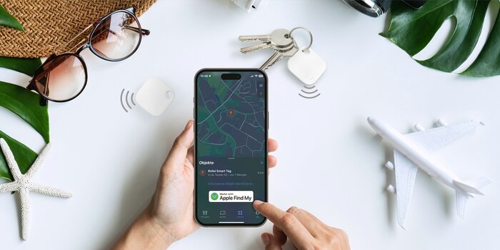 Draufsicht. Hänge halten ein Smartphone mit geöffneter Wo-ist-App. Auf dem Tisch liegt ein Schlüsselbung mit dem Rollei Smart-Tag sowie Urlaubsdekoration Draufsicht. Hänge halten ein Smartphone mit geöffneter Wo-ist-App. Auf dem Tisch liegt ein Schlüsselbung mit dem Rollei Smart-Tag sowie Urlaubsdekoration