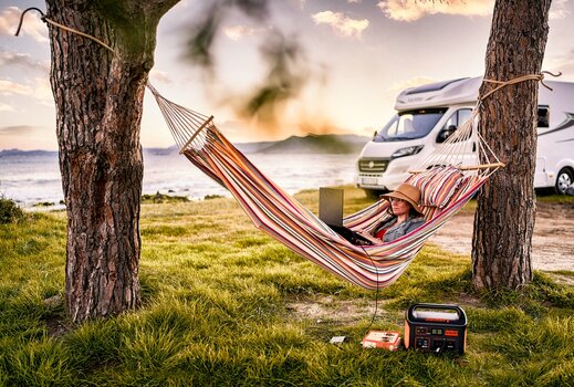Frau liegt in Hängematte und arbeitet am Laptop, der mit einem Jackery Explorer mit Strom versorgt wird. Im Hintergrund Wohnmobil und Küstenlandschaft.