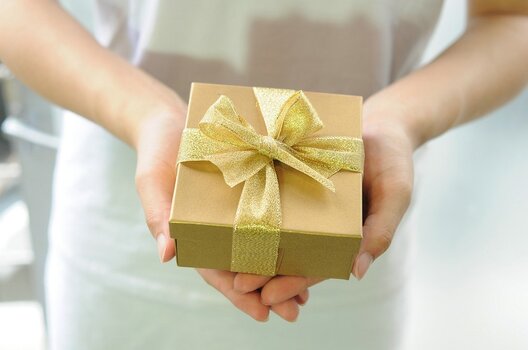 Frau im weißen Kleid hält eine goldene Geschenkverpackung in beiden Händen in die Kamera. Symbolbild.