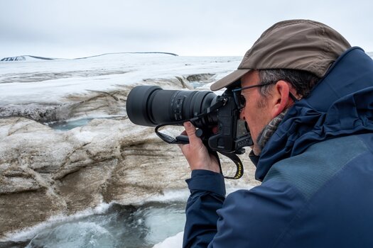 Mann in blauer Jacke und mit brauner Mütze am rechten untreren Bildrand, hält eine Kamera mit Tele-Objektiv in den Händen und vor sein Gesicht, schaut durch den Sucher, als Motiv dient ihm eine eisige Steinlandschaft, Arktis
