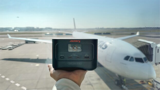 Jackery Mini-Powerstation Explorer 100 Plus auf ausgestreckter Handfläche. Im Hintergrund ist ein Flugzeug auf einem Flughafen zu sehen.