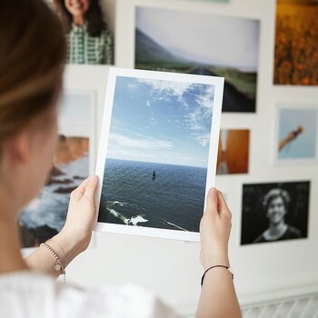 Eine junge Frau hält einen hochwertigen Fotodruck einer Meereslandschaft mit einem Segelboot in den Händen. Im Hintergrund hängen weitere ausgedruckte Bilder an einer hellen Wand, was die emotionale Wirkung gedruckter Fotos unterstreicht.