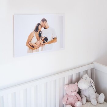 Ein großformatiger Leinwanddruck eines emotionalen Familienporträts hängt über einem weißen Kinderbett. Das Bild zeigt die Bedeutung von gedruckten Erinnerungen als langlebige und persönliche Wanddekoration im Wohnbereich.