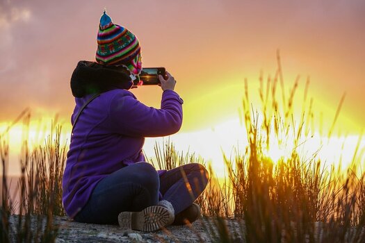 Eine Person sitzt im Schneidersitz im hohen Gras und fotografiert mit einem Smartphone einen leuchtenden Sonnenuntergang. Sie trägt eine lila Fleecejacke und eine bunte, gemusterte Strickmütze mit Ohrenklappen.