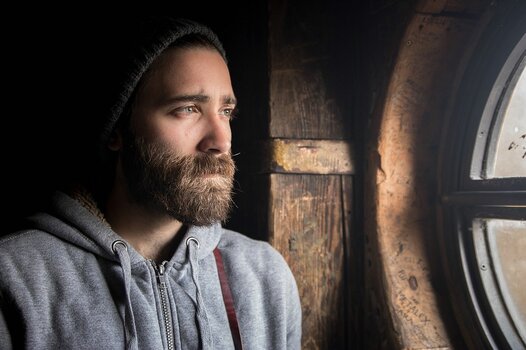 Porträt eines bärtigen Mannes mit dunkler Strickmütze und grauem Kapuzenpullover. Er blickt nachdenklich aus einem runden, rustikalen Holzfenster. Das Licht fällt von draußen auf sein Gesicht, während der Innenraum im Schatten liegt. Porträt eines bärtigen Mannes mit dunkler Strickmütze und grauem Kapuzenpullover. Er blickt nachdenklich aus einem runden, rustikalen Holzfenster. Das Licht fällt von draußen auf sein Gesicht, während der Innenraum im Schatten liegt.