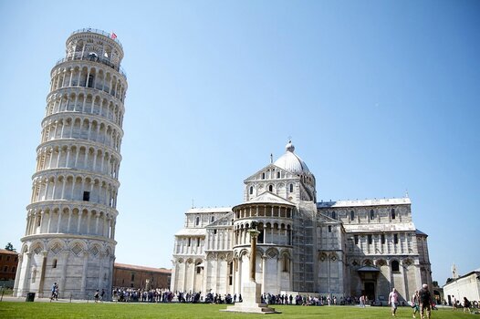 Der Schiefe Turm von Pisa und die angrenzende Kathedrale bei strahlendem Sonnenschein mit zahlreichen Touristen im Vordergrund.