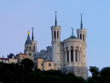 Die weiße Basilika Notre-Dame de Fourvière in Lyon auf einem bewaldeten Hügel vor hellem Abendhimmel.