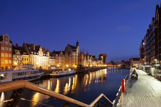 Historische Häuserfronten am Ufer der Mottlau in Danzig während der Blauen Stunde mit hell leuchtenden Straßenlaternen und Lichtreflexionen im Wasser.