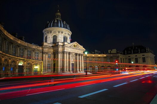 Langzeitbelichtung des Institut de France in Paris bei Nacht, bei der vorbeifahrende Autos leuchtend rote und weiße Lichtspuren auf der Straße im Vordergrund hinterlassen.