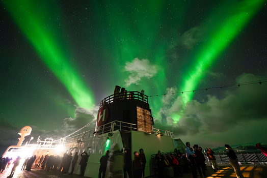 Passagiere an Deck eines Hurtigruten-Schiffes unter einem strahlend grünen Polarlicht-Himmel während der Fahrt durch Nordnorwegen.